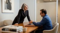 Professional blonde businesswoman in black suit consulting with male colleague in modern office meeting room for corporate strategy discussion.
