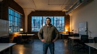 Middle-aged Caucasian man standing confidently in modern industrial loft office space with exposed brick walls, large windows and contemporary furniture.