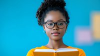 Young African American woman with natural curly hair and glasses reading book against blue background, expressing focus and intelligence. Professional portrait.