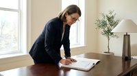 Professional businesswoman in dark suit reviewing documents at wooden desk in bright modern office with natural lighting for corporate presentations.