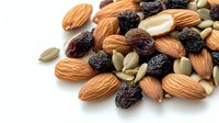 Healthy mix of raw almonds, dark raisins and pumpkin seeds scattered on white background, macro food photography showing natural texture and details.