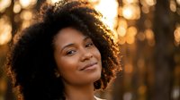 Young African American woman with curly hair smiling warmly in golden hour sunlight with bokeh background for lifestyle and beauty content.