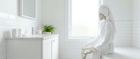 Woman in white bathrobe and towel sitting on toilet in bright, minimalist bathroom with subway tile walls and white vanity sink.