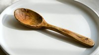 Handcrafted wooden spoon with natural grain pattern on white ceramic plate, rustic kitchen utensil for cooking and serving organic food preparation.