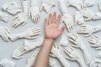 Living hand reaching out among multiple white plaster cast hands arranged on light gray background, symbolizing uniqueness, individuality and standing out from crowd.