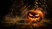 Glowing carved pumpkin with menacing face sits on ground surrounded by dried vines and brick wall, emitting eerie orange light for seasonal decorations.