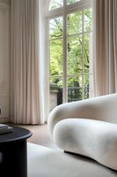 Modern curved white sofa near tall window with flowing curtains and green garden view. Natural light streams into minimalist luxury interior space.