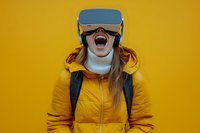 Young woman in yellow winter jacket wearing VR headset laughing with excitement against vibrant yellow background, expressing joy and amazement in virtual reality.