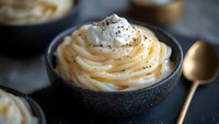 Creamy butterscotch pudding topped with whipped cream and spices served in dark ceramic bowl with gold spoon, selective focus on swirled dessert surface.