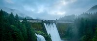 Hydroelectric dam with powerful water spillway surrounded by misty forest mountains during overcast weather conditions for renewable energy concepts.