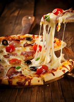 Hot pizza slice with melted cheese stretching, topped with pepperoni, olives and tomatoes on rustic wooden table background for restaurant menu design.