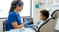 Female dentist in blue scrubs showing dental X-rays to young mixed-race boy patient in modern clinic chair during pediatric consultation appointment.