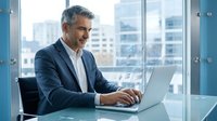 Mature businessman with gray hair smiling while working on laptop in modern office with city view. Professional executive in suit typing at desk.