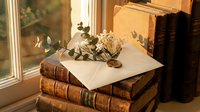 Vintage leather-bound books stacked by sunny window with dried eucalyptus branch and envelope, creating warm nostalgic reading atmosphere for literary themes.