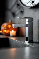 Modern chrome bathroom faucet on dark countertop with Halloween decorations and spider silhouette in blurred background.