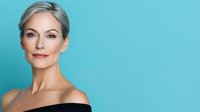 Elegant mature woman with silver hair and natural makeup smiling confidently against turquoise background, showcasing beauty and aging gracefully. Professional portrait for beauty and wellness industry.