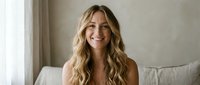 Happy young blonde woman with wavy hair smiling warmly while sitting on bed in bright modern bedroom interior for lifestyle and wellness content.