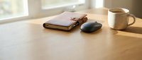 Minimalist workspace with leather notebook, wireless mouse and coffee mug on wooden desk near window. Clean office setup for productivity and focus.