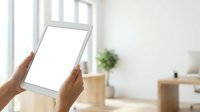 Close-up of hands holding blank tablet with white screen in bright modern office workspace with natural light and wooden desk accessories.
