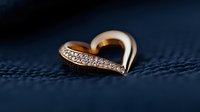 Golden heart-shaped pendant with sparkling diamonds on dark blue textured fabric background, macro jewelry photography for luxury accessories and romance concepts.