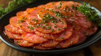 Thinly sliced cured meat carpaccio garnished with fresh herbs and black pepper on dark plate, served as gourmet appetizer on wooden surface.