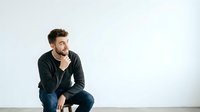 Thoughtful man with beard in casual dark hoodie sitting and contemplating against white background for lifestyle and portrait use.