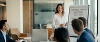 Professional businesswoman presenting strategic growth initiatives to corporate team in modern conference room with whiteboard and charts.
