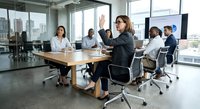 Professional businesswoman raising hand during corporate meeting in modern glass conference room with diverse team of colleagues seated around wooden table.