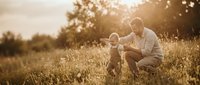 Father and young child bonding in golden hour meadow with warm sunlight creating magical bokeh effect through tall grass and trees in peaceful countryside setting.