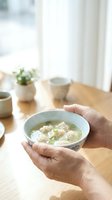 Person holding bowl of wonton soup with green onions at wooden table near window, cups and potted plant in background for food and dining concepts.