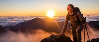 Female hiker in outdoor gear ascending mountain peak at sunrise above clouds with trekking poles, adventure and exploration concept.