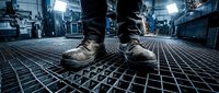 Worker in heavy-duty boots stands on industrial metal grating floor in factory or warehouse setting with blue lighting and machinery in background.