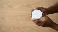 Close-up of dark-skinned hands holding blank white round container against wooden surface. Suitable for product packaging, cosmetics, skincare, and branding mockups.
