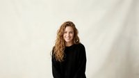 Young Caucasian woman with curly blonde hair wearing black sweater smiling at camera against neutral beige wall background in natural lighting.