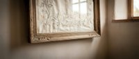 Elegant ornate golden picture frame hanging on beige wall in soft natural light from window creating warm atmospheric interior decor scene.