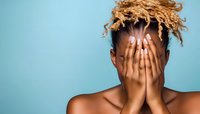 Young African American woman with blonde curly hair covering face with hands against turquoise background, expressing emotion and vulnerability.