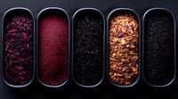 Assorted dried tea varieties in dark ceramic containers showing different textures and colors from purple petals to red powder and black loose leaf tea.