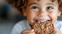 Toddler with curly reddish-brown hair and light skin tone biting into a whole grain cookie, showing joyful expression. Suitable for parenting, nutrition, and child wellness content.