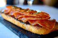 Crispy baguette topped with thinly sliced prosciutto and fresh herbs served on dark stone plate, close up view of gourmet appetizer with selective focus.