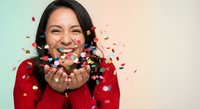 Happy young Hispanic woman in red sweater blowing colorful confetti from hands celebrating holiday season with joyful expression against pastel background.
