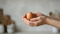Caucasian woman's hands holding a brown chicken egg against a blurred kitchen background, representing fresh organic produce, healthy eating, cooking ingredients, and farm-to-table food preparation.