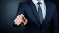 Businessman in dark suit pointing finger forward against black background, conveying leadership and direct communication in professional setting.