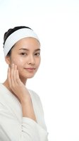 Young Asian woman applying facial cream with white headband touching cheek in skincare routine on white background for beauty and wellness concepts.