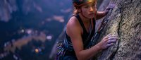 Young athletic woman rock climbing on granite cliff face during golden hour sunset with safety gear and harness for outdoor adventure sports.