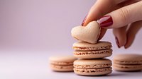 Female hand with red manicure holding heart shaped macaron cookie over pink macarons background, romantic food photography for Valentine's day and love concept.
