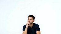 Man with dark hair and olive complexion wearing navy blue t-shirt, hand to face in thoughtful pose, isolated on white background for design and marketing use.