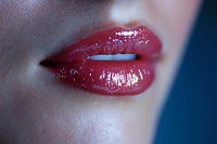 Close-up of glossy red lips with wet effect makeup against dark blue background, showing professional cosmetic application and sensual expression.