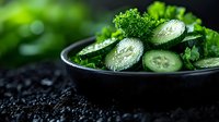 Fresh sliced cucumber and parsley in black ceramic bowl on dark background with water drops, macro food photography for healthy eating concept.