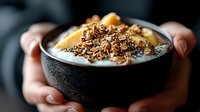 Creamy yogurt bowl topped with crunchy homemade granola, honey drizzle and nuts in black ceramic bowl held by hands. Dark moody breakfast photography.