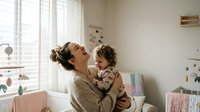 Young mother with messy bun holding laughing toddler son in bright modern nursery room with natural light from window creating warm family moment.